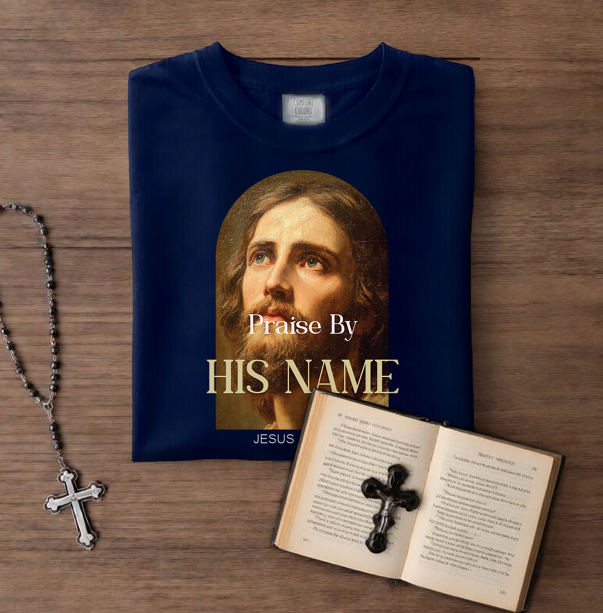 Navy blue t-shirt with Jesus face and text on a wooden surface with a rosary and open book.