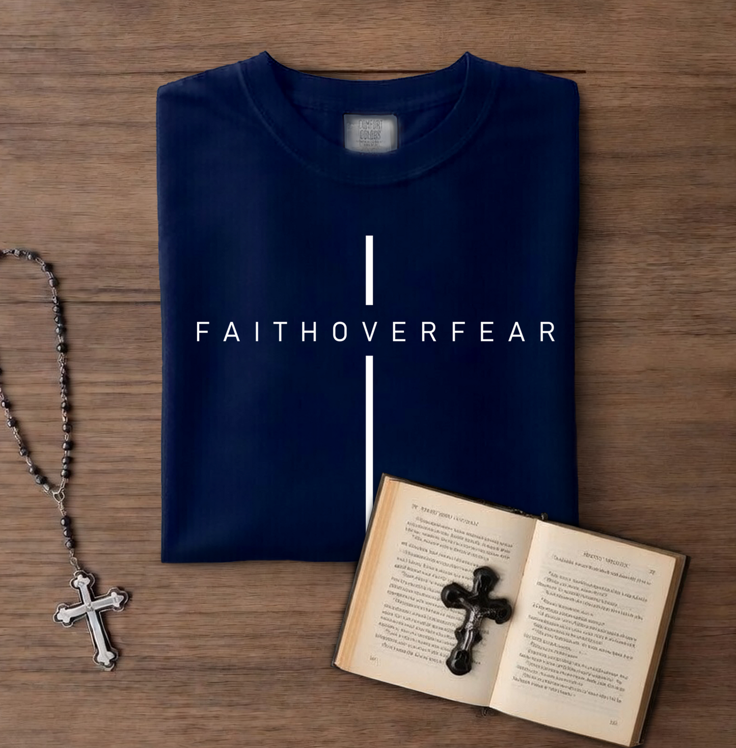 Folded navy blue shirt with 'FAITHOVERFEAR' text, rosary, and open book with cross on wooden surface