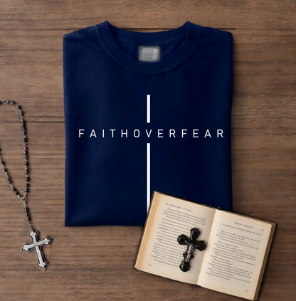 Folded navy blue shirt with 'FAITHOVERFEAR' text, rosary, and open book with cross on wooden surface