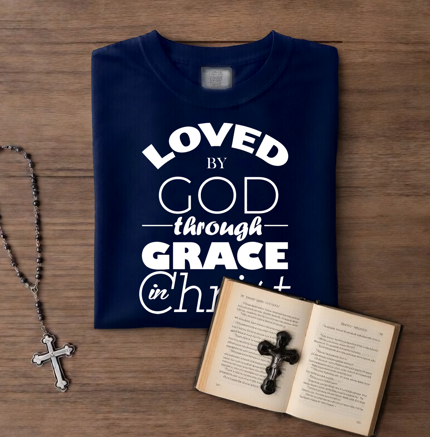 Navy blue t-shirt with 'LOVED by GOD through GRACE in CHRIST' text on a wooden surface with a book and rosary.