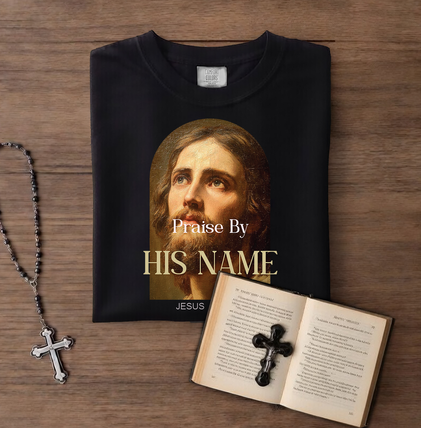 Black t-shirt with a portrait of Jesus and 'Praise By HIS NAME' text, next to a rosary and open book with a cross on a wooden surface.