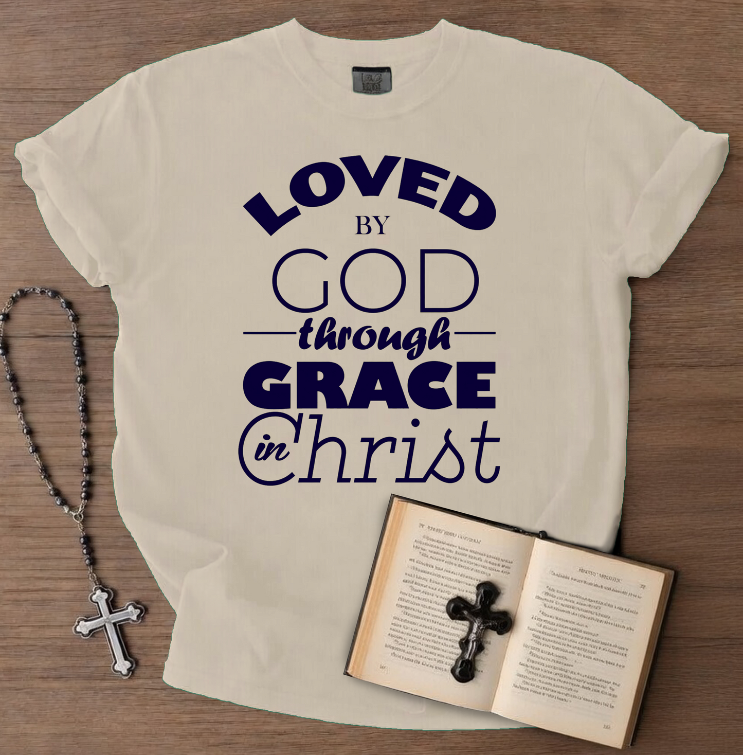 Beige t-shirt with 'Loved by God through Grace in Christ' text, next to a rosary and open book on a wooden surface.