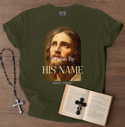 Green t-shirt with a graphic of a religious figure and text, placed on a wooden surface with a book and rosary.