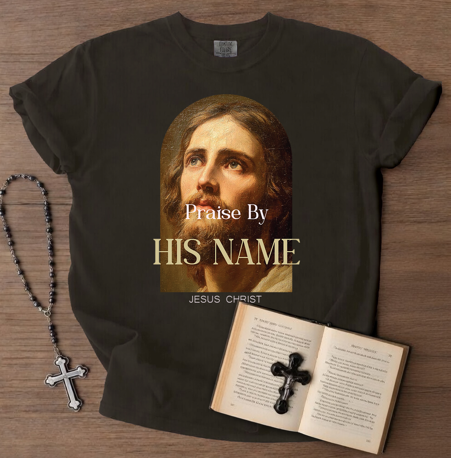 Black t-shirt with a graphic of Jesus Christ and text 'Praise By HIS NAME JESUS CHRIST' on a wooden surface, with a rosary and open book with a cross.