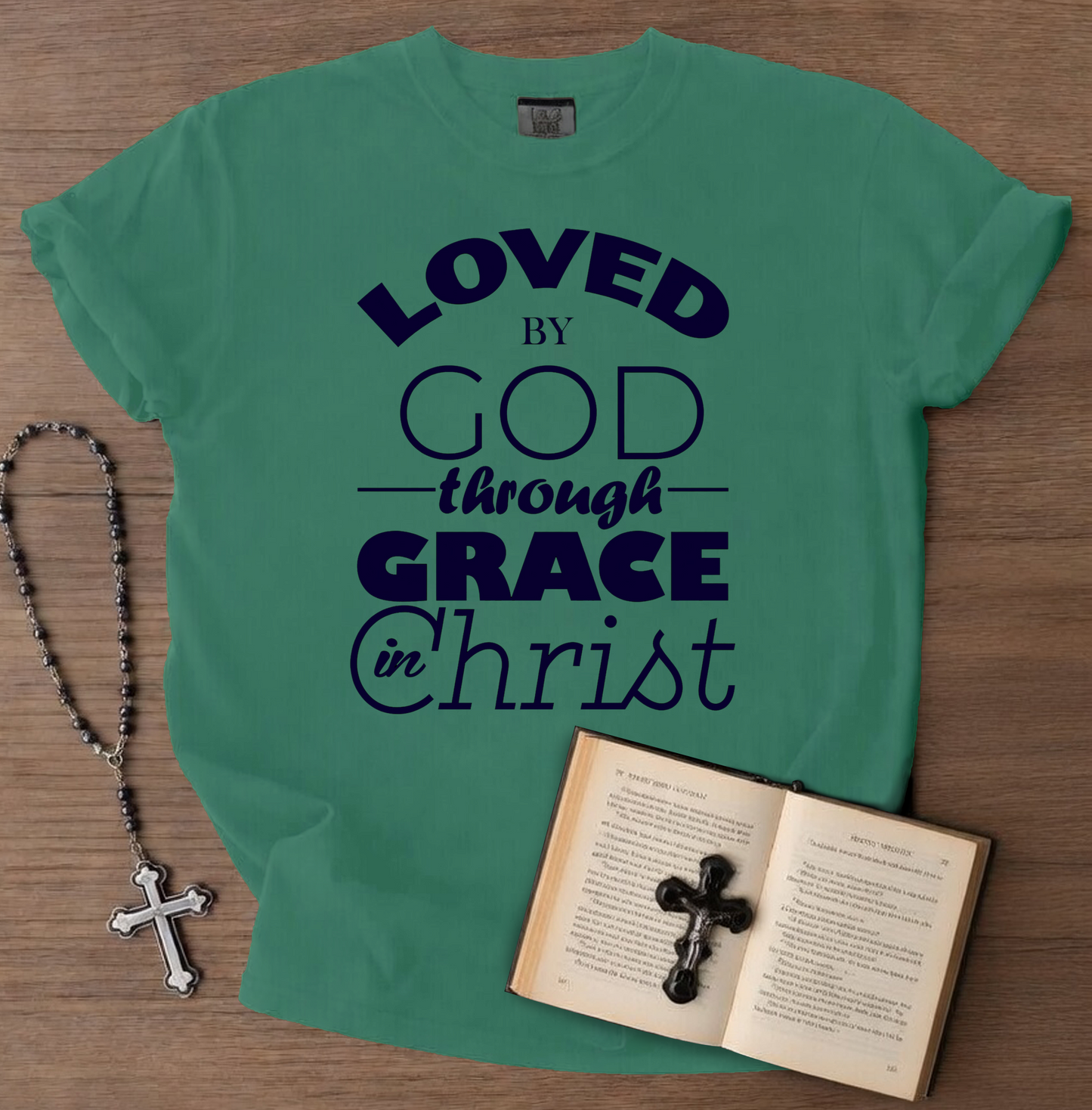 Green t-shirt with 'Loved by God through Grace in Christ' text, next to a book and rosary on a wooden surface.