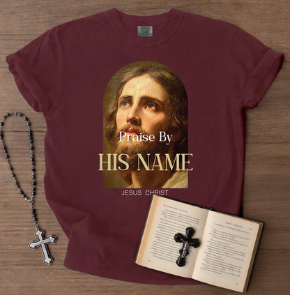 Maroon t-shirt with Jesus Christ graphic and text on a wooden surface with a book and rosary.