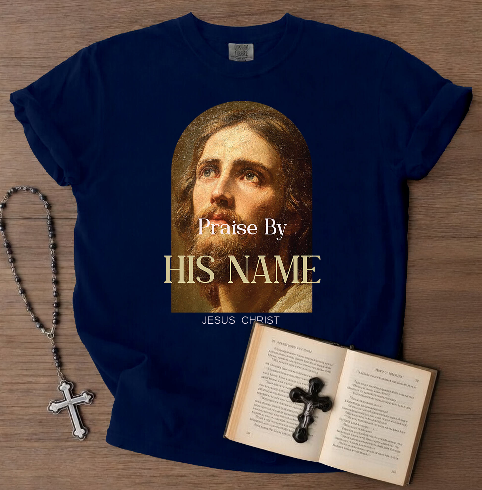 Navy blue t-shirt with a graphic of a religious figure and text, placed on a wooden surface with a rosary and open book.