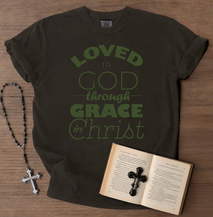 Black t-shirt with green text 'LOVED by GOD through GRACE in Christ' on a wooden surface with a book, cross, and rosary.