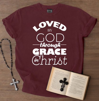 Maroon t-shirt with 'Loved by God through Grace in Christ' text, next to a rosary and open book on a wooden surface.
