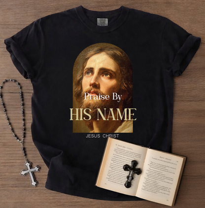 Black t-shirt with a graphic of Jesus Christ and text 'Praise By HIS NAME JESUS CHRIST' on a wooden surface, accompanied by a rosary and open book with a cross.