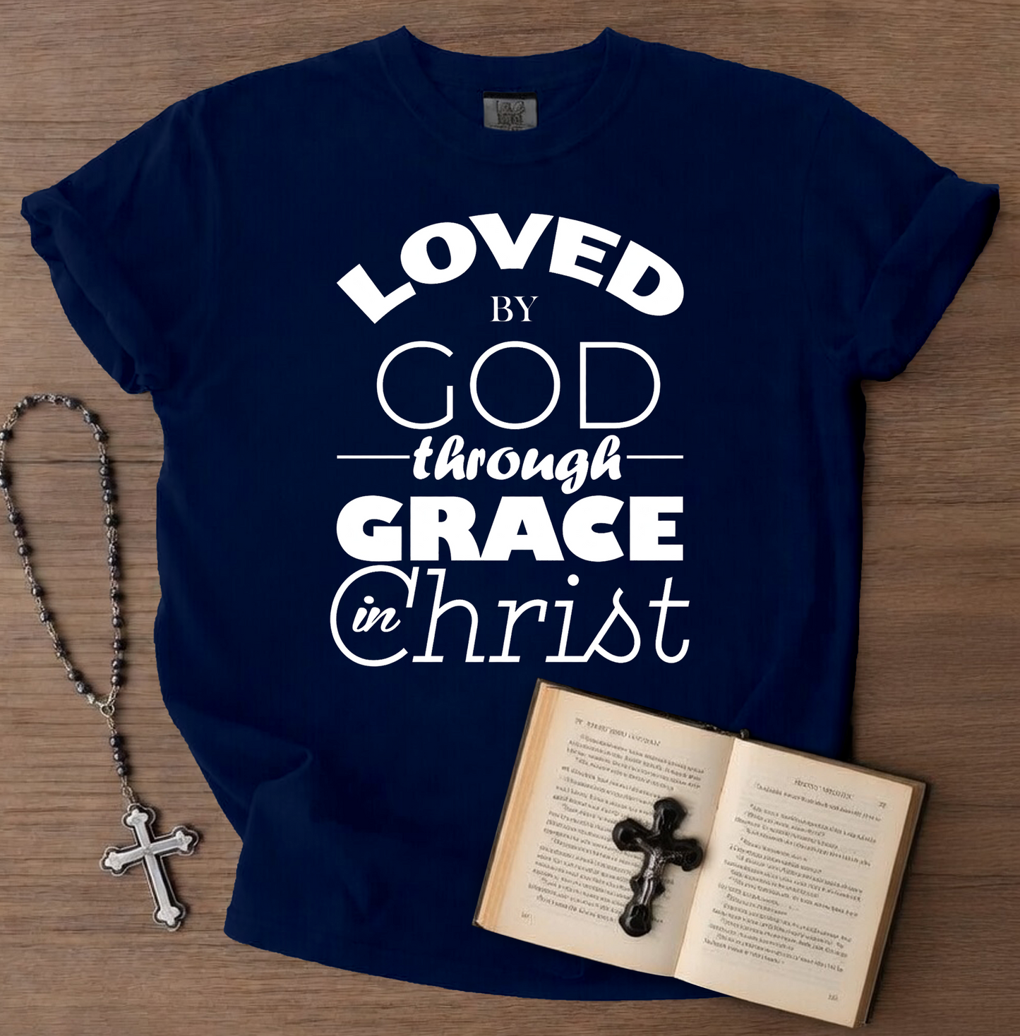 Navy blue t-shirt with 'LOVED by GOD through GRACE in Christ' text, placed on a wooden surface with a book and rosary.