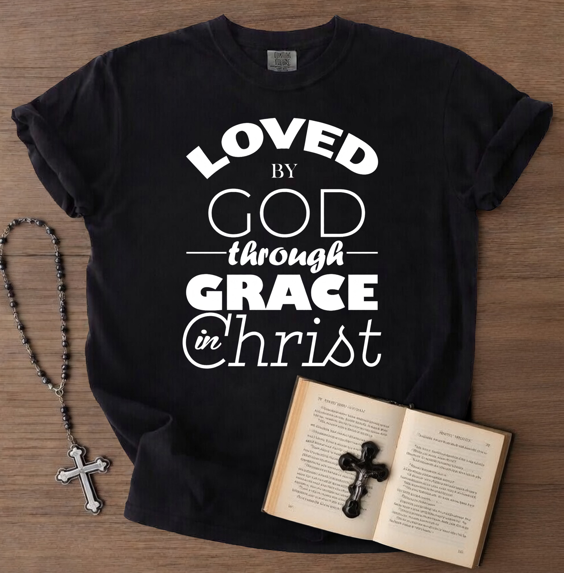 Black t-shirt with 'Loved by God through Grace in Christ' text, next to an open book with a cross and a rosary on a wooden surface.