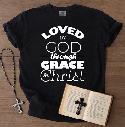 Black t-shirt with 'Loved by God through Grace in Christ' text, next to an open book with a cross and a rosary on a wooden surface.