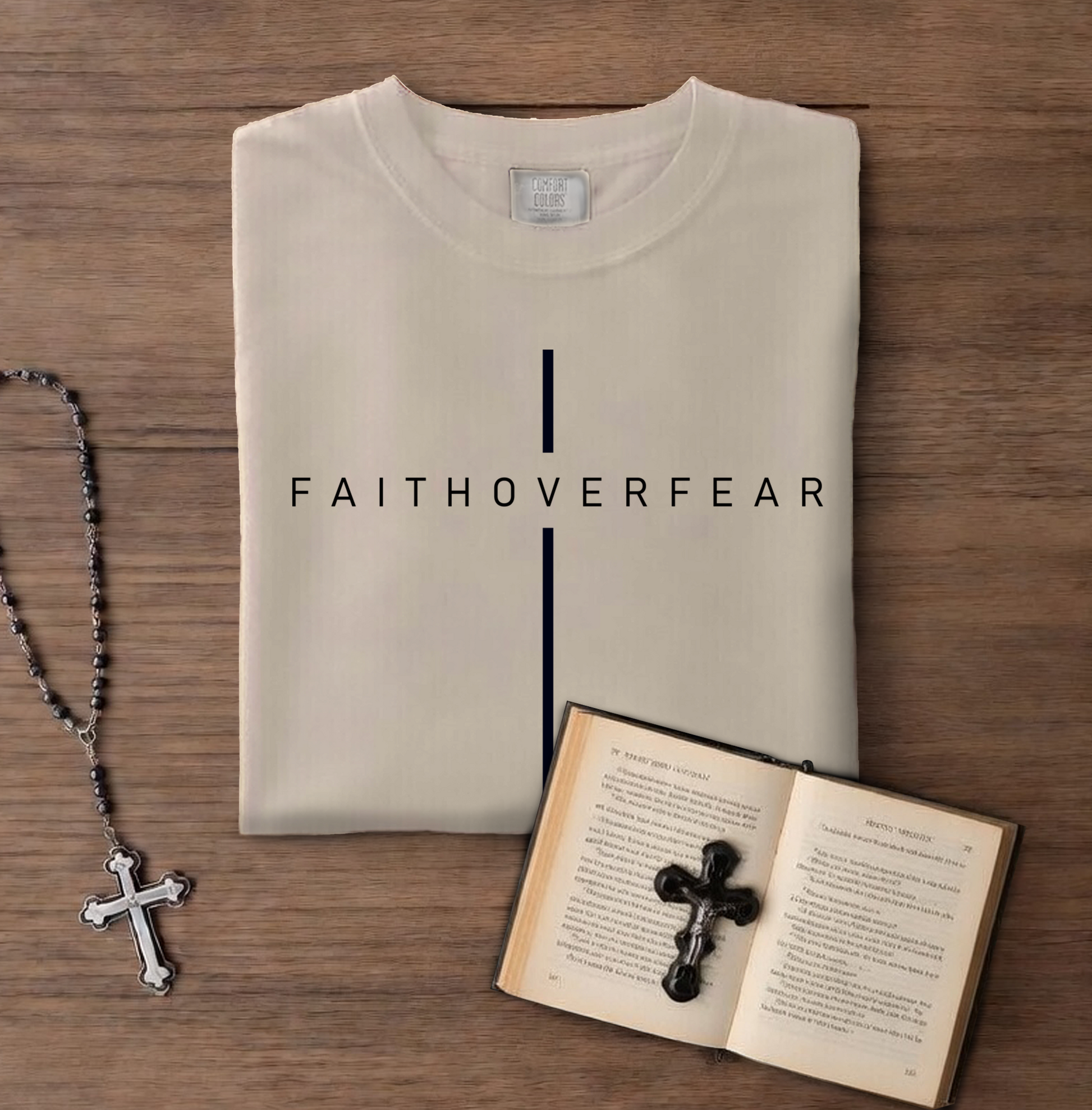 Folded t-shirt with 'FAITHOVERFEAR' text, rosary, and open book on wooden surface