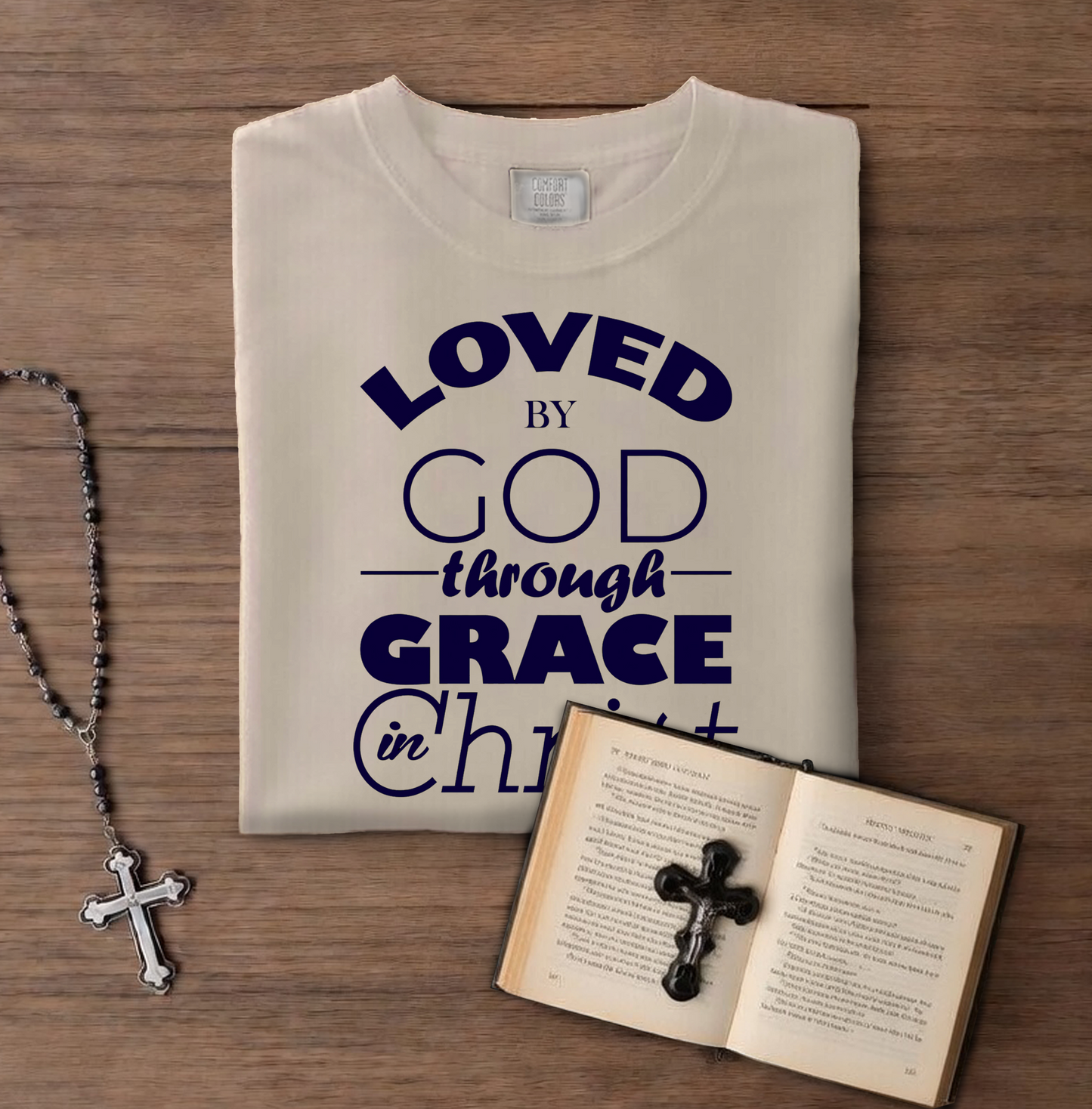 Folded t-shirt with 'LOVED BY GOD through GRACE in CHRIST' text on a wooden surface with a book and cross necklace.