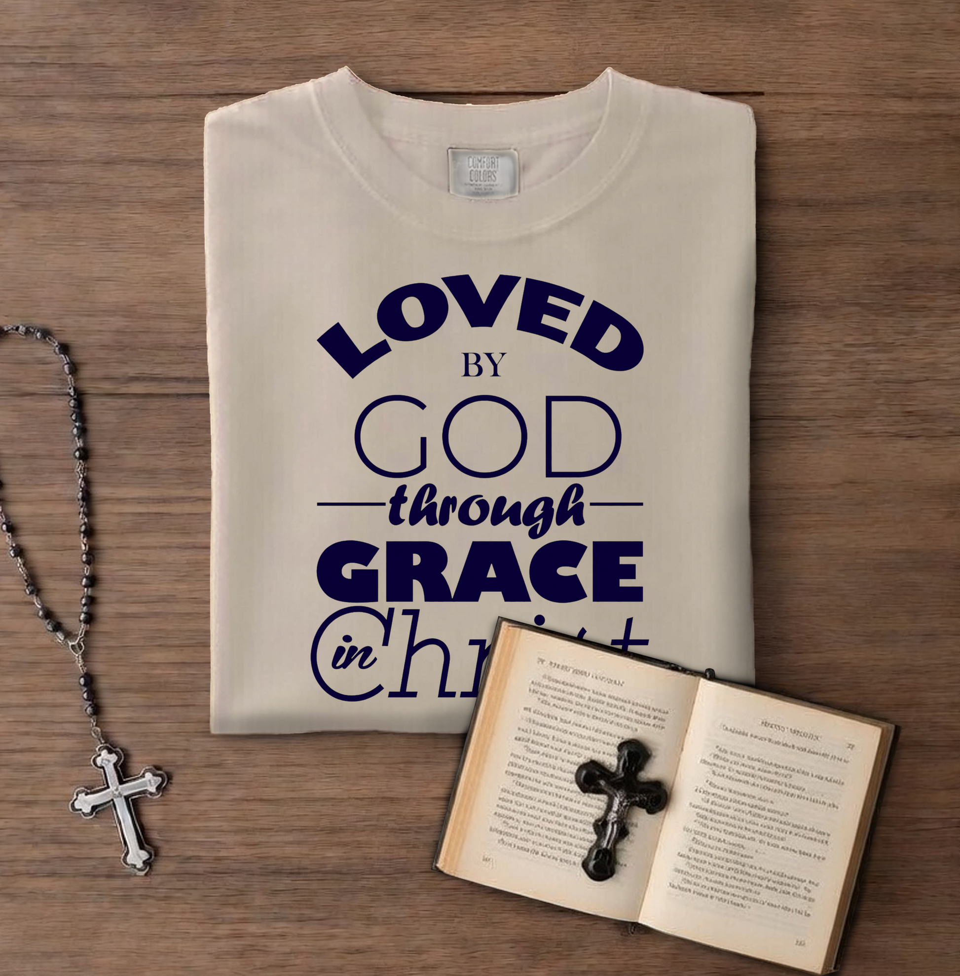 Folded t-shirt with 'LOVED BY GOD through GRACE in CHRIST' text on a wooden surface with a book and cross necklace.