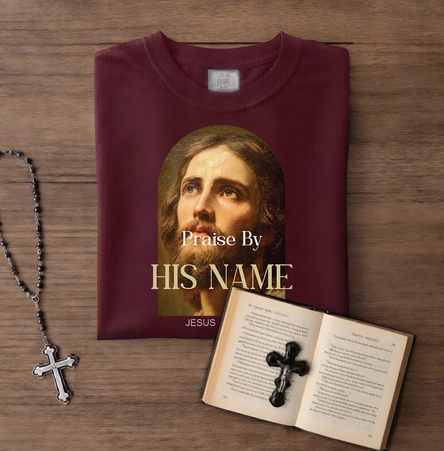 Maroon t-shirt with Jesus' face and 'Praise By HIS NAME JESUS' text on a wooden surface, next to a rosary and open book with a cross.