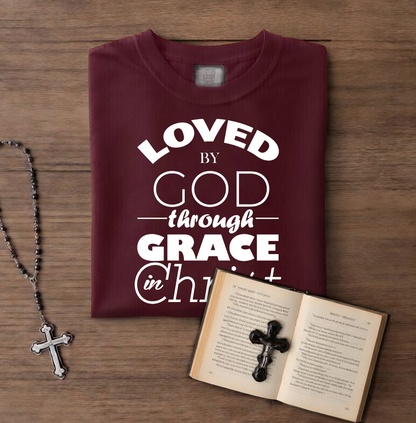 Maroon t-shirt with 'Loved by God through Grace in Christ' text on a wooden surface with a book and rosary.