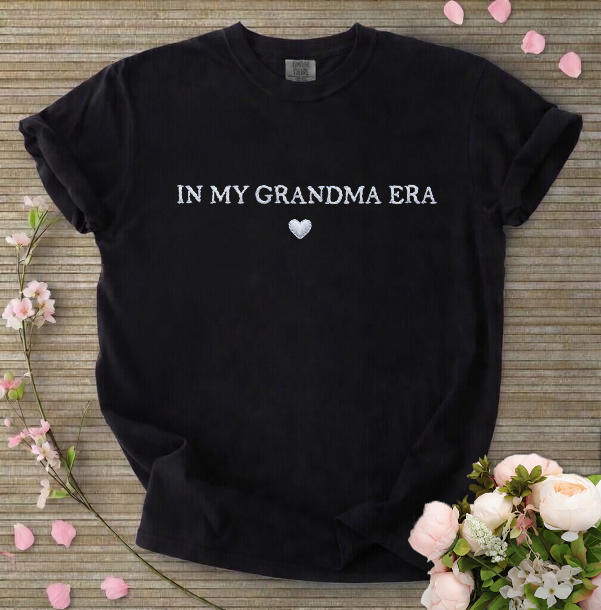 Black t-shirt with 'IN MY GRANDMA ERA' text on a wooden surface with flowers.