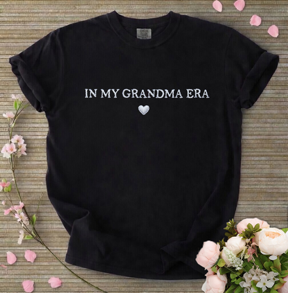 Black t-shirt with 'IN MY GRANDMA ERA' text on a wooden surface with flowers.