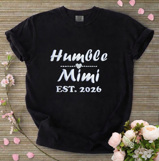Black t-shirt with 'Humble Mimi EST. 2026' text on a wooden surface with flowers.