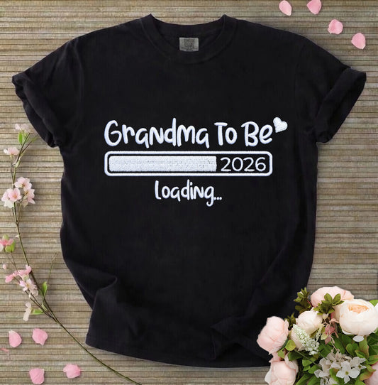 Black t-shirt with 'Grandma To Be 2026 loading...' text on a wooden surface with flowers.