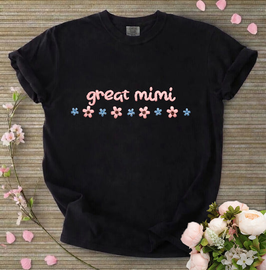 Black t-shirt with 'great mimi' text and flowers on a wooden surface with flowers around