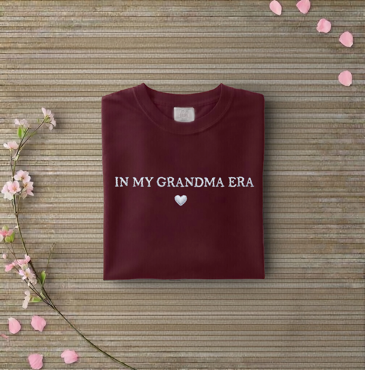 Maroon sweatshirt with 'IN MY GRANDMA ERA' text on a wooden surface with flowers and petals.