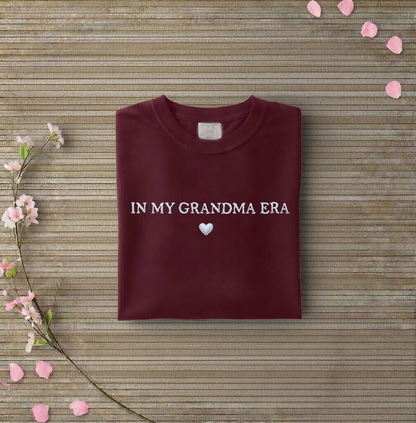 Maroon sweatshirt with 'IN MY GRANDMA ERA' text on a wooden surface with flowers and petals.