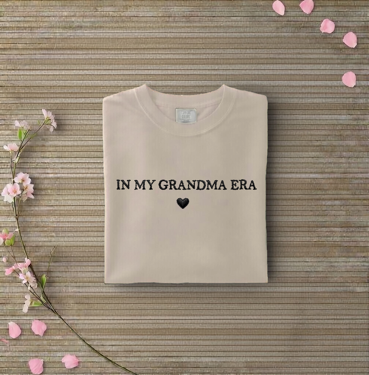 Beige sweatshirt with 'IN MY GRANDMA ERA' text on a wooden surface with flowers and petals.