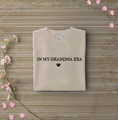 Beige sweatshirt with 'IN MY GRANDMA ERA' text on a wooden surface with flowers and petals.