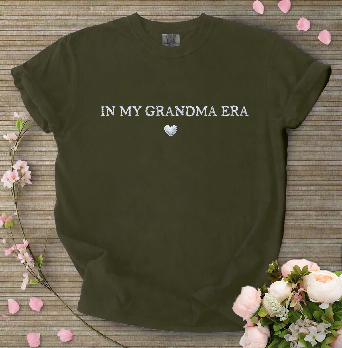 Dark green t-shirt with 'IN MY GRANDMA ERA' text on a wooden surface with flowers.