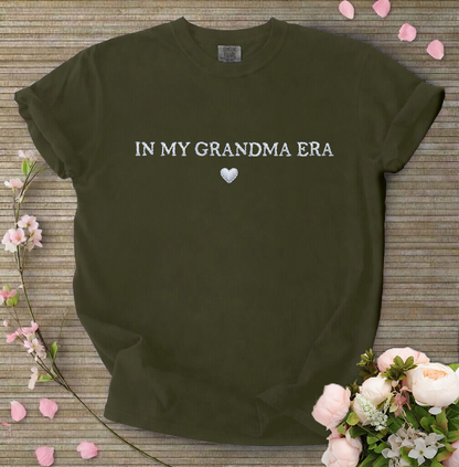 Dark green t-shirt with 'IN MY GRANDMA ERA' text on a wooden surface with flowers.