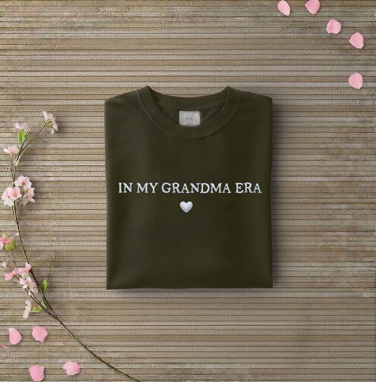 Dark green sweatshirt with 'IN MY GRANDMA ERA' text on a wooden surface with flowers and petals.