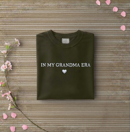 Dark green sweatshirt with 'IN MY GRANDMA ERA' text on a wooden surface with flowers and petals.