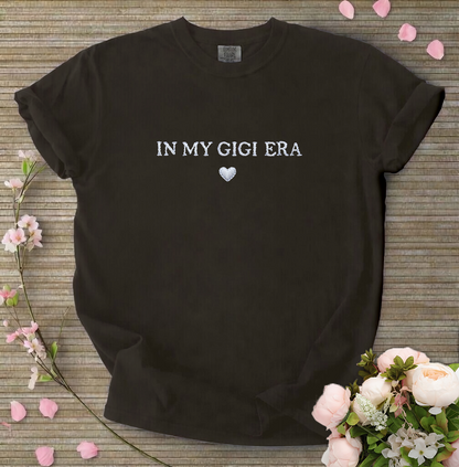Black t-shirt with 'IN MY GIGI ERA' text on a wooden surface with flowers.