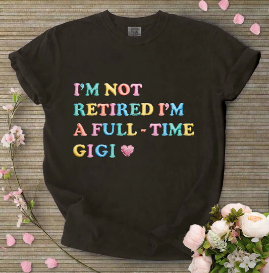 Black t-shirt with colorful text 'I'M NOT RETIRED I'M A FULL-TIME GIGI' on a wooden surface with flowers.