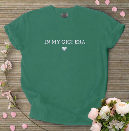 Green t-shirt with 'IN MY GIGI ERA' text on a wooden surface with flowers.