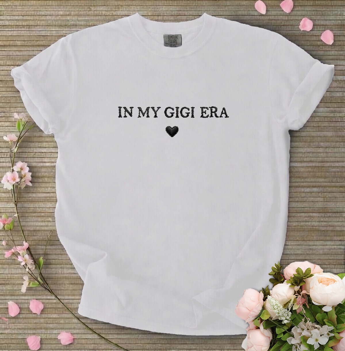 White t-shirt with 'IN MY GIGI ERA' text on a wooden surface with flowers and petals.